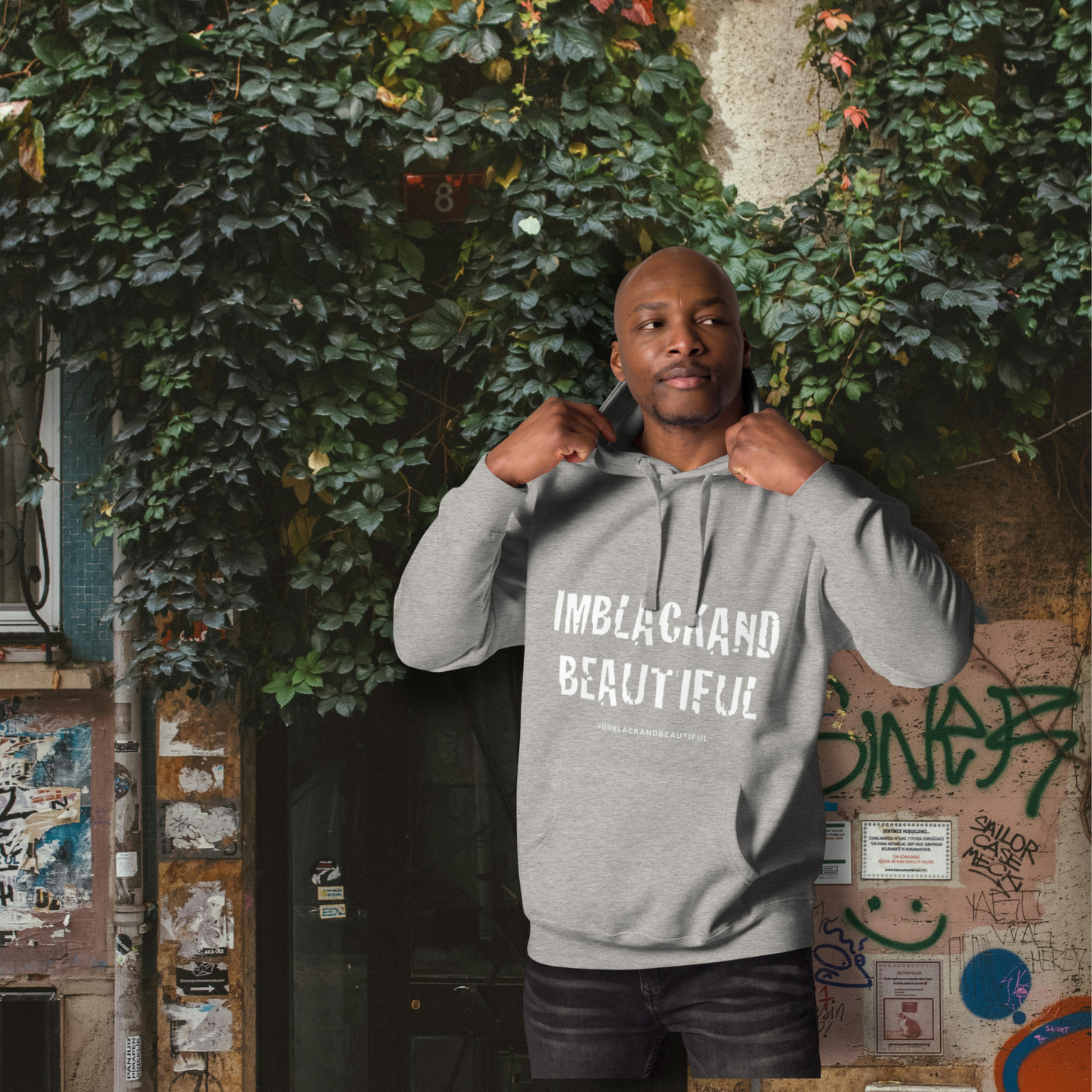 Man wearing a gray hoodie with text, standing in front of a wall with greenery and graffiti.