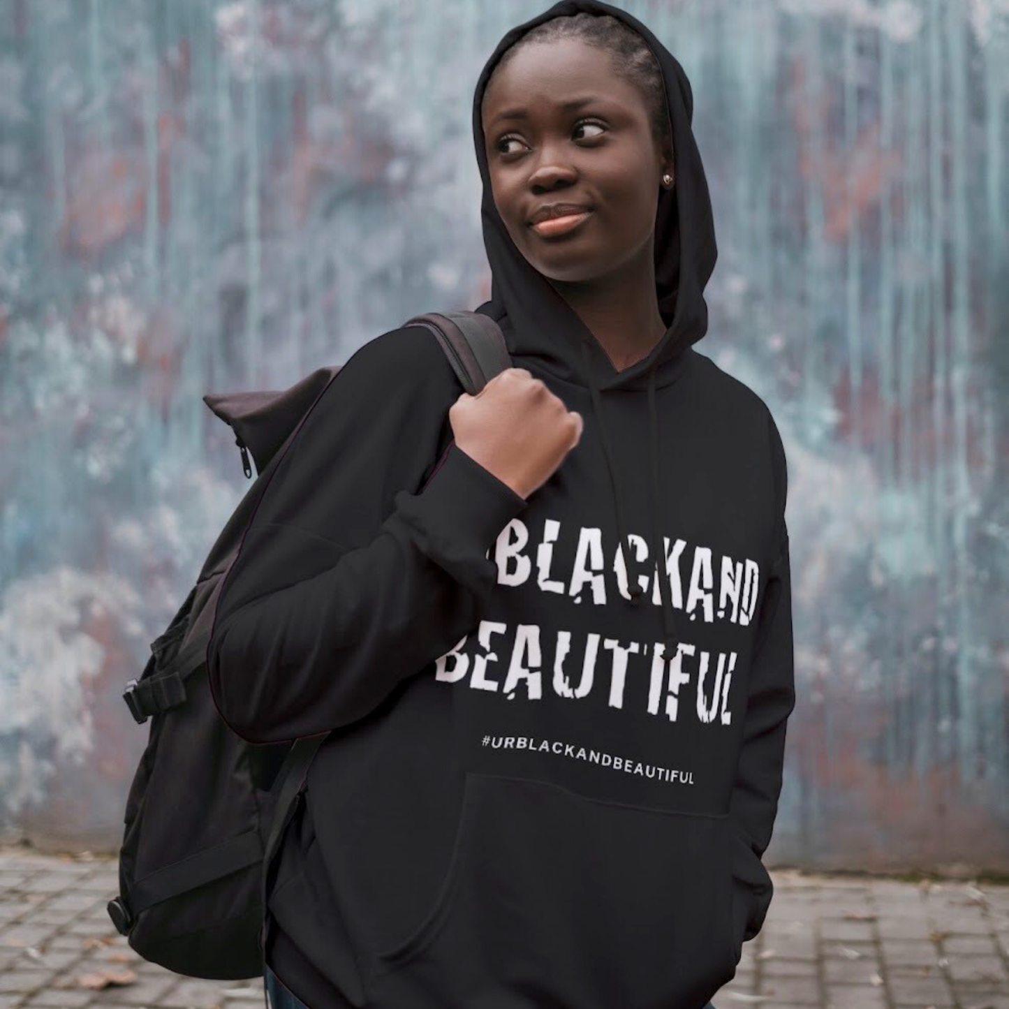 Person wearing a black hoodie with 'BLACK AND BEAUTIFUL' text, holding a backpack against a textured wall.